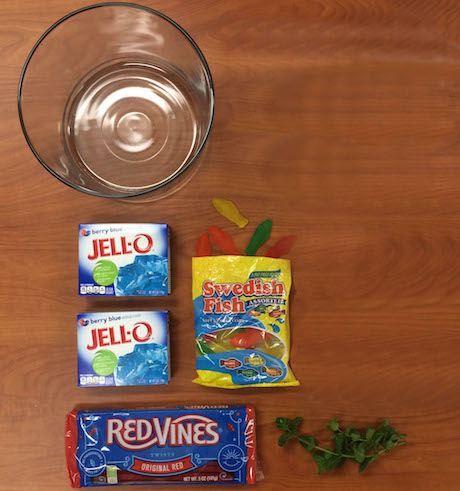 ingredients on table, such as red licorice, blue gelatin dessert, glass bowl, gummy fish